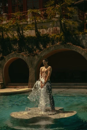 Shirtless man stands in swimming pool on stone platform with water flowing from his hands, with architectural backdrop and trees.