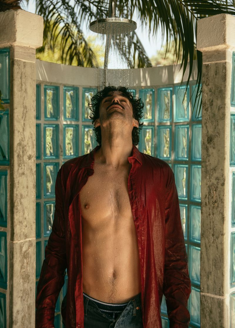 Man standing in outdoor shower with shirt on. Shower water cascading down over body. Tropical greenery in background, sunny day.
