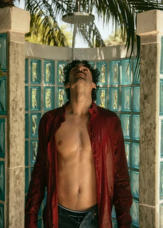 Man standing in outdoor shower with shirt on. Shower water cascading down over body. Tropical greenery in background, sunny day.