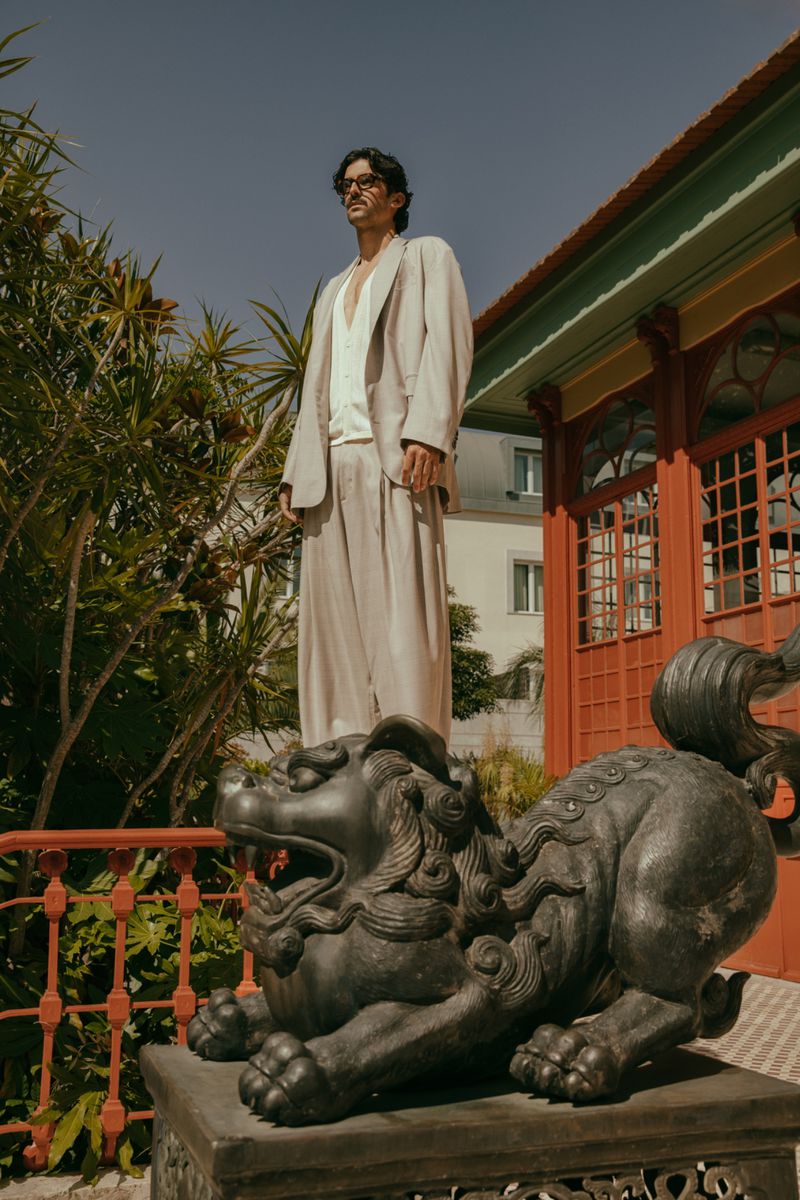Man in a light beige suit standing on a lion statue outside a red building surrounded by greenery, beneath a clear blue sky.