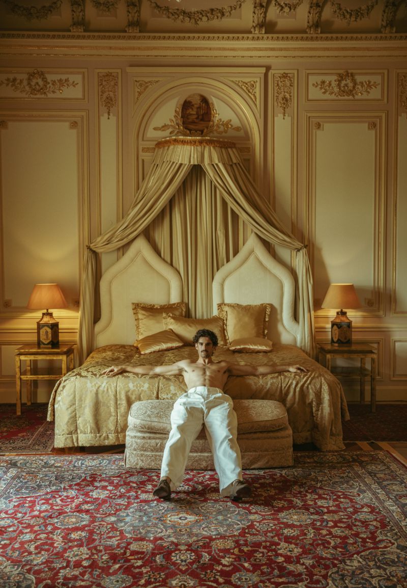 A man in white pants sits on a cushioned bench in front of a luxurious bed inside an ornate room with pale walls and detailed mouldings.