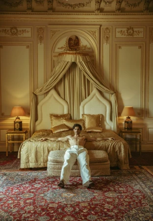 A man in white pants sits on a cushioned bench in front of a luxurious bed inside an ornate room with pale walls and detailed mouldings.