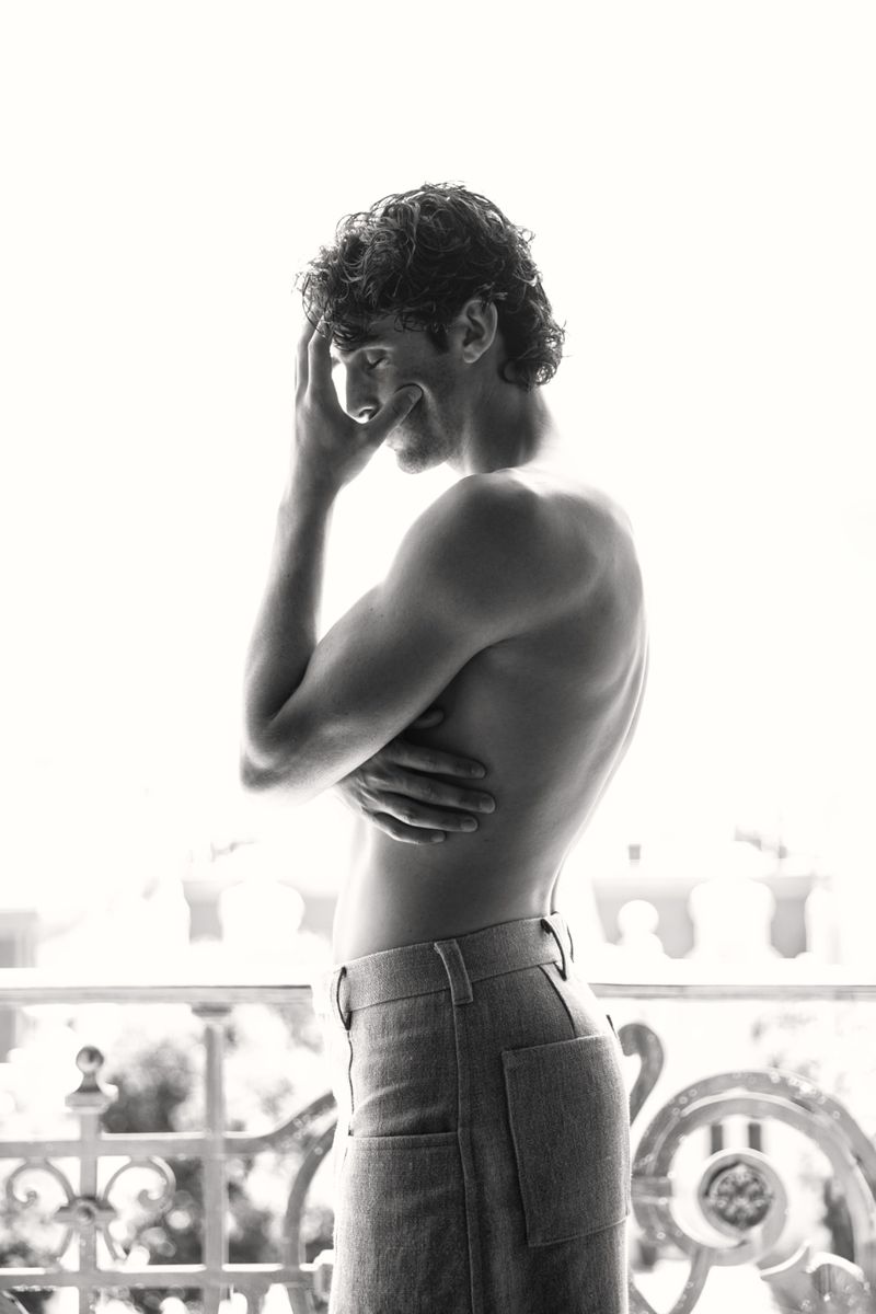 Black and white portrait of a fit adult male with curly hair, turned away from the camera, wearing jeans, standing near an iron balcony.