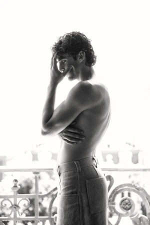 Black and white portrait of a fit adult male with curly hair, turned away from the camera, wearing jeans, standing near an iron balcony.