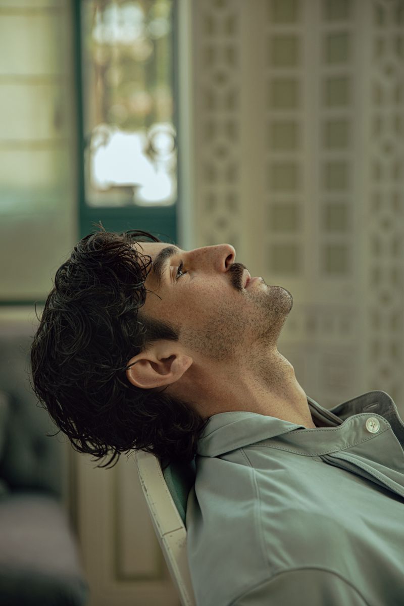 Portrait of a dark-haired man with a mustache leaning back in a chair in a room with soft, diffused natural lighting indoors.