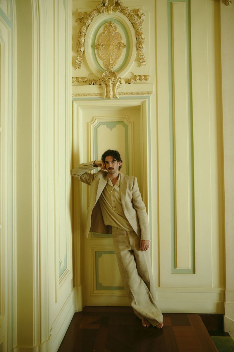 Man poses in beige suit leaning against decorative wall and doorway in an ornate interior with hardwood floor and pale yellow walls.