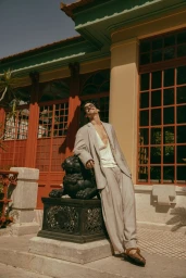 Man in a relaxed light grey suit leaning against a decorative black lion statue outside a building with red window frames.