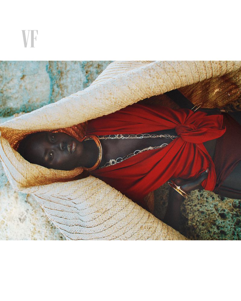 Dark-skinned woman with a straw hat and red fabric, accessorized with necklaces and bracelets, lying on a textured surface.