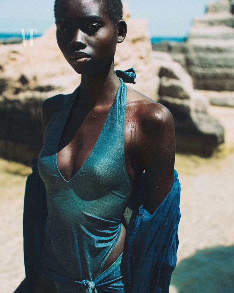 Close-up shot of an African woman in a teal halter top and wrap standing on a beach with rock formations and light-blue ocean in the background.