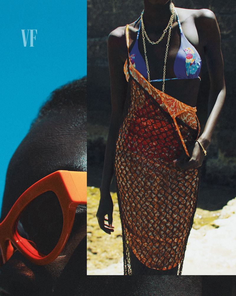 Fashion editorial shot of a dark-skinned female model in a bikini top, gold jewelry and fishnet dress posing outdoors on a sunny day.
