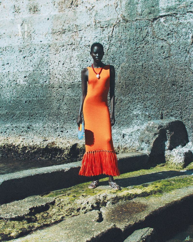 Fashion model in orange fringe dress posing outdoors against a concrete wall and steps. Accessorized with orange necklace and handbag.