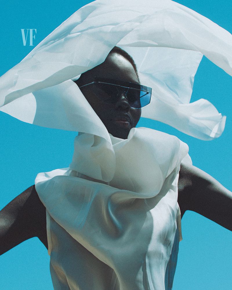 Elegant portrait of a black woman in sunglasses and white flowing top against a blue sky with white translucent fabric overhead.