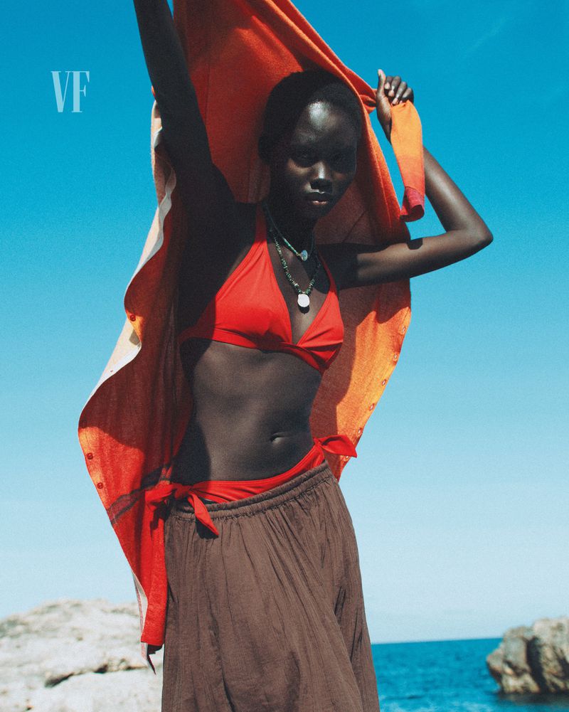 Woman in red bikini top and brown skirt holding orange textile on a rocky beach against a blue sky and ocean background.