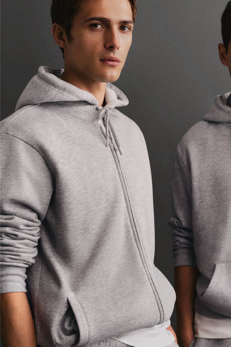 Close-up of a young adult male wearing a gray zip-up hoodie, standing indoors against a grey wall, in a fashion or lifestyle product shot.