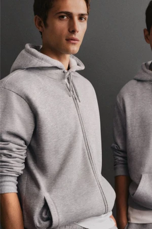 Close-up of a young adult male wearing a gray zip-up hoodie, standing indoors against a grey wall, in a fashion or lifestyle product shot.