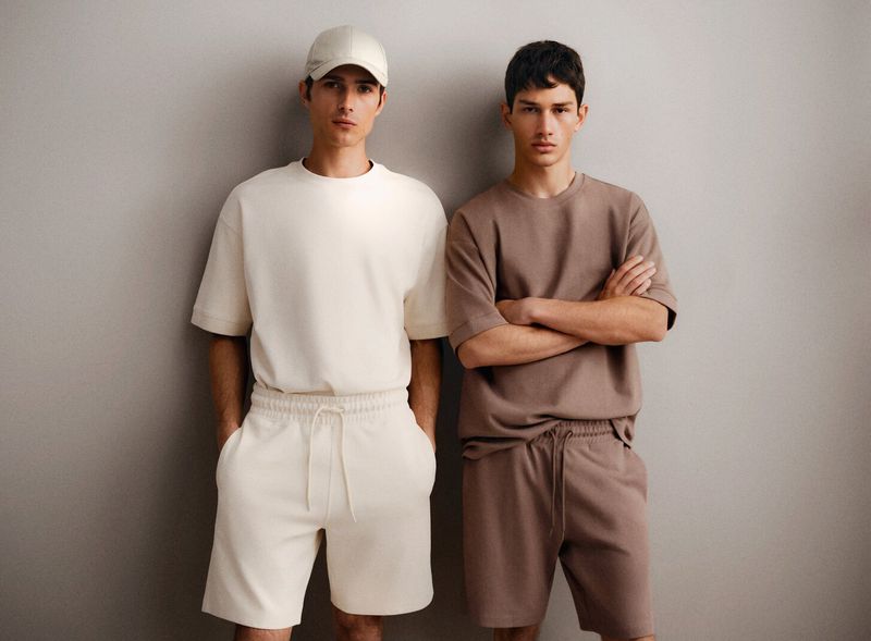 Two young adult men pose in matching casual outfits of t-shirts and shorts in a studio with soft lighting against a neutral gray background.