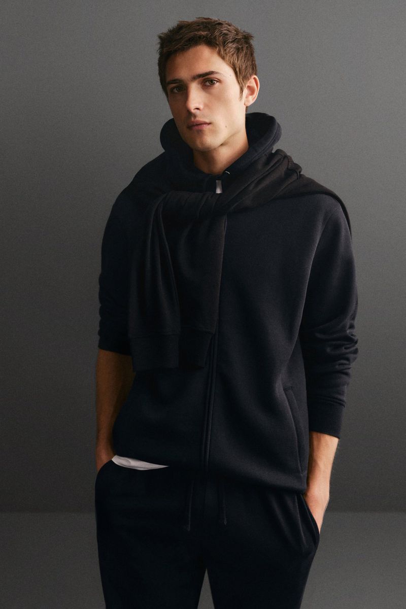 Adult male poses indoors wearing a black zip-up hoodie and sweatpants, with a sweater draped over his shoulders against a grey backdrop.