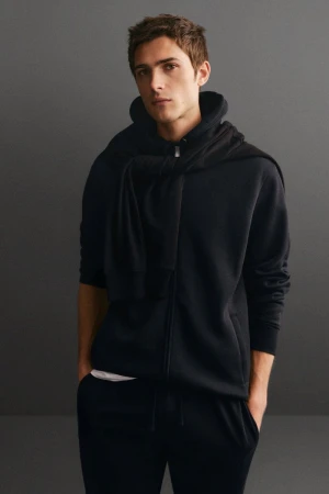 Adult male poses indoors wearing a black zip-up hoodie and sweatpants, with a sweater draped over his shoulders against a grey backdrop.