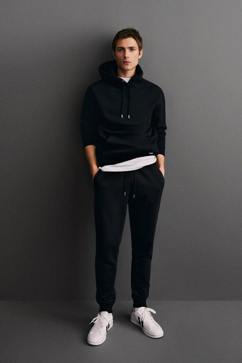 Full-length portrait of a young adult male in a black hooded sweatshirt and sweatpants, standing against a solid grey background.