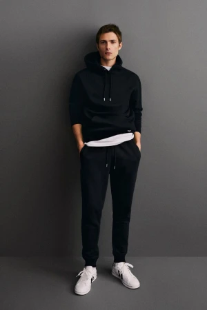 Full-length portrait of a young adult male in a black hooded sweatshirt and sweatpants, standing against a solid grey background.