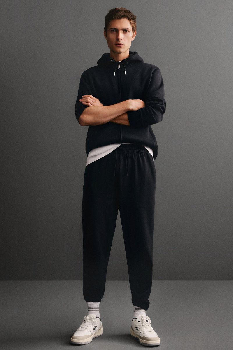 Full shot of a young adult male with crossed arms, wearing a black zip-up hoodie and sweatpants with white sneakers against a gray backdrop.