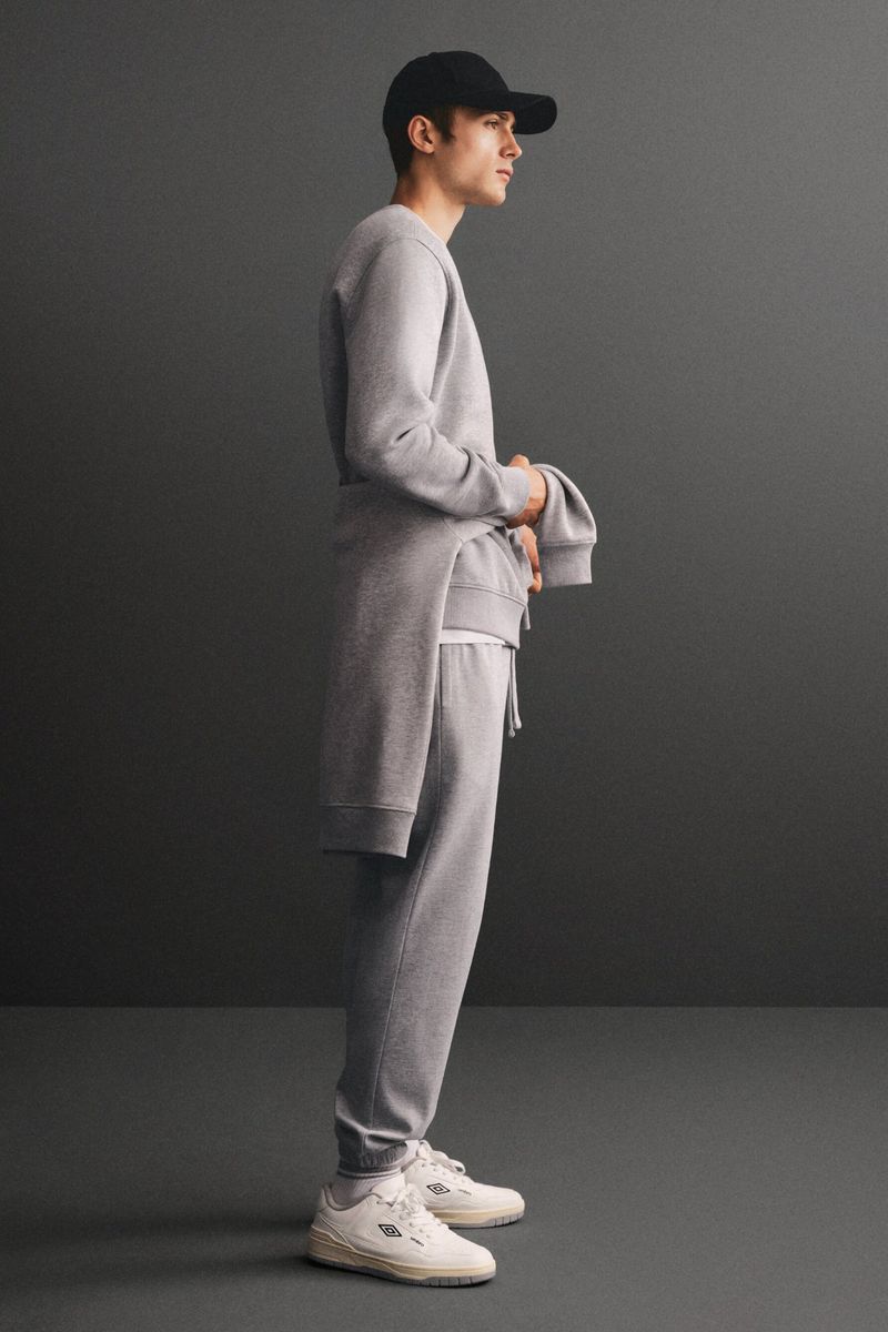 Full body shot of a young adult man in casual grey sweat suit and black baseball cap posing in a grey studio, fashion advertising.