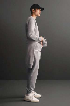 Full body shot of a young adult man in casual grey sweat suit and black baseball cap posing in a grey studio, fashion advertising.