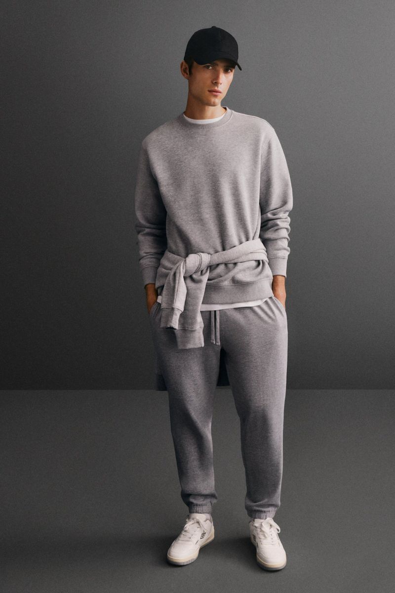 An adult male in a grey sweatshirt and sweatpants poses indoors in front of a grey wall, wearing a black baseball cap and white sneakers.