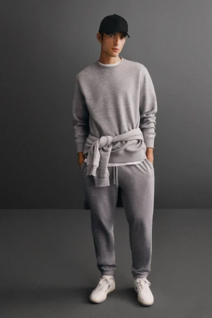 An adult male in a grey sweatshirt and sweatpants poses indoors in front of a grey wall, wearing a black baseball cap and white sneakers.