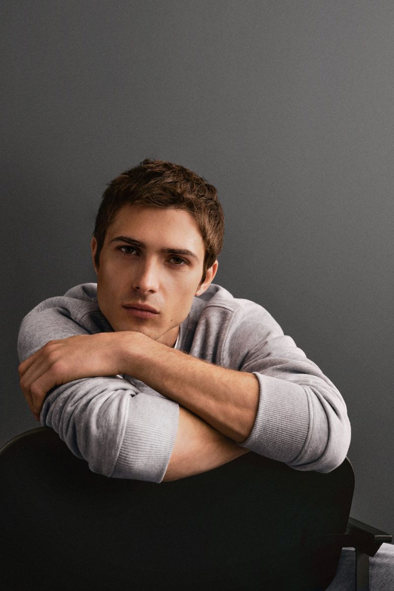 Portrait of a young adult male with short brown hair, wearing a gray sweatshirt, leaning on the back of a black chair against gray background.