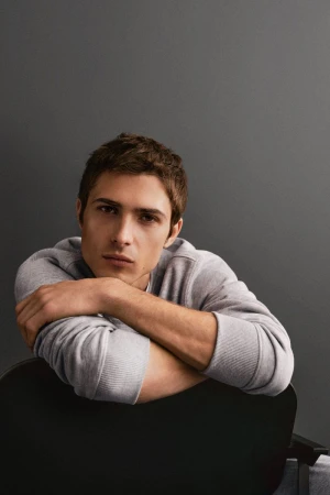 Portrait of a young adult male with short brown hair, wearing a gray sweatshirt, leaning on the back of a black chair against gray background.