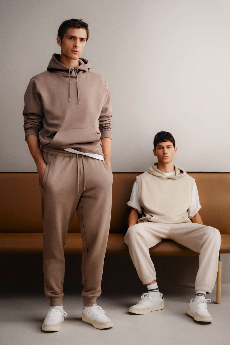 Two adult males pose indoors wearing matching casual lounge wear of hoodies, sweatpants, and sneakers in a fashion lifestyle shot.