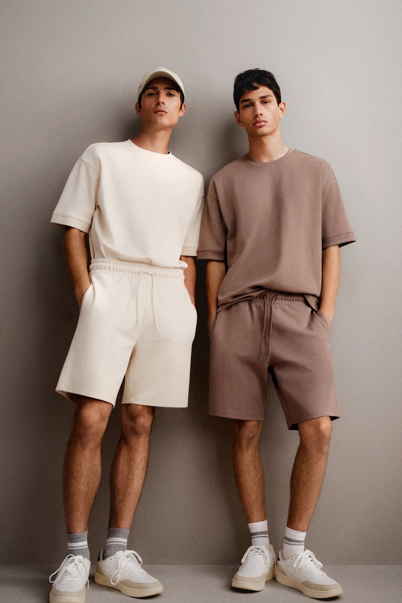 Two young men pose indoors wearing matching casual outfits: short-sleeved shirts, shorts, socks, and sneakers in a fashion editorial.