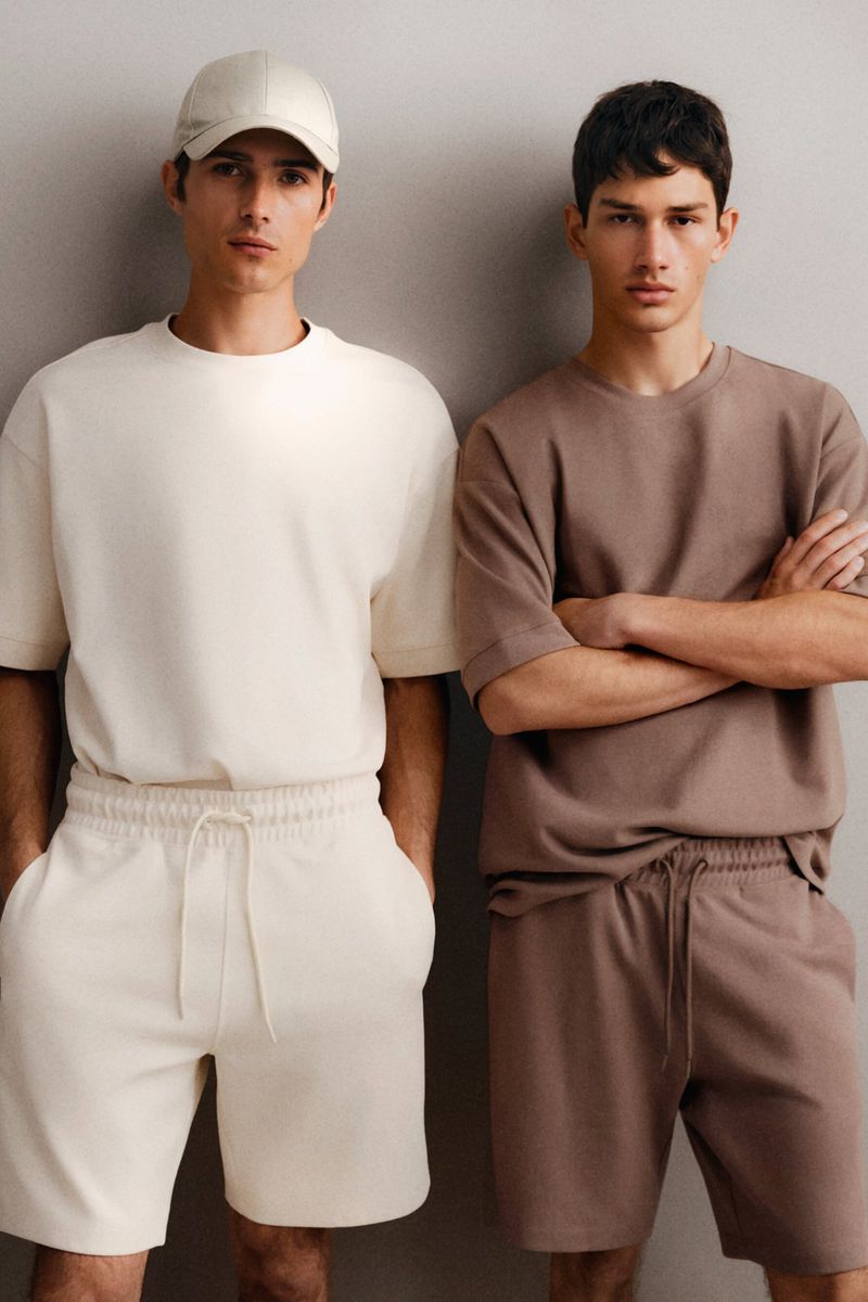 Two adult men pose in matching knit t-shirts and shorts in a studio. One man wears a baseball cap and the other has arms crossed.