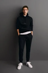 A male model wearing a black hoodie and sweatpants with white sneakers against a neutral grey studio background.