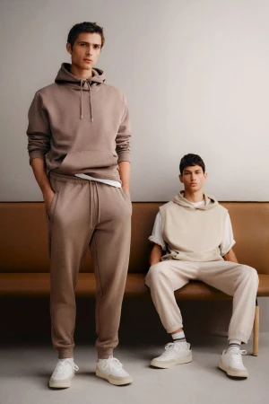 Two male models in neutral-toned hoodies and sweatpants posing against a plain grey studio backdrop with a leather bench