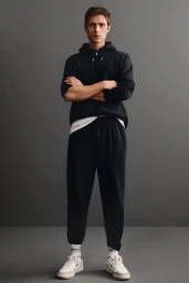 Full-body shot of a male model wearing a black zip hoodie, black sweatpants, and white sneakers against a plain grey background.