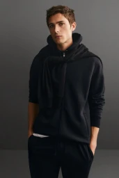 A male model wearing a black hoodie and matching trousers posing against a dark grey studio background.