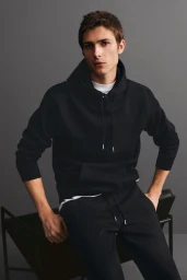 A young male model in a black hoodie and black sweatpants leaning against a dark chair in front of a grey wall.