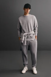 Model wearing a grey sweatshirt, matching sweatpants, white sneakers, and a black baseball cap against a dark grey background