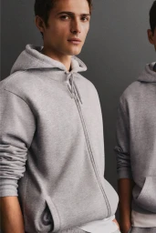 Close-up of a young man wearing a light grey zip-up hoodie against a neutral dark grey studio backdrop.