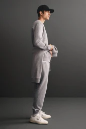 Profile shot of a young man wearing a grey sweatshirt, grey sweatpants, and a black cap against a dark studio background