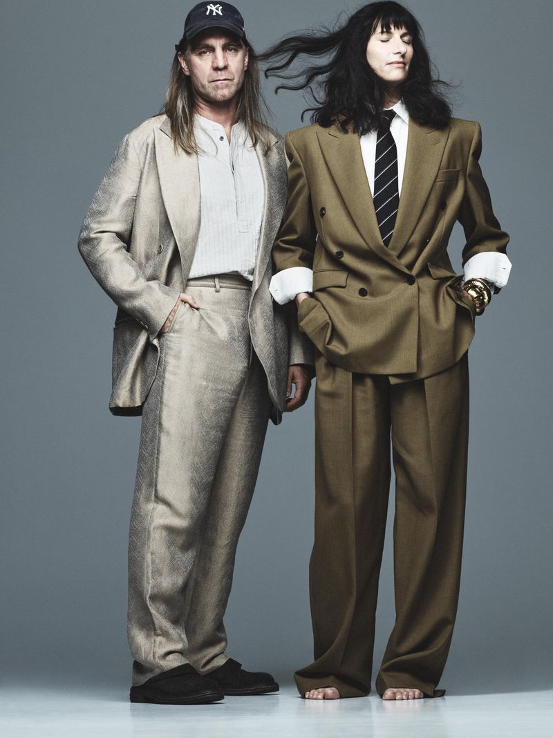 Two people pose in tailored suits, one in light tan, the other in olive, against a neutral backdrop in a fashion editorial.