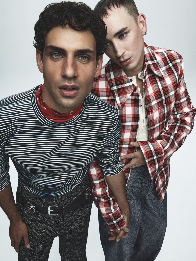 Two men pose in a brightly lit studio, one in a striped shirt and the other in a red and white plaid shirt, holding hands, portrait.