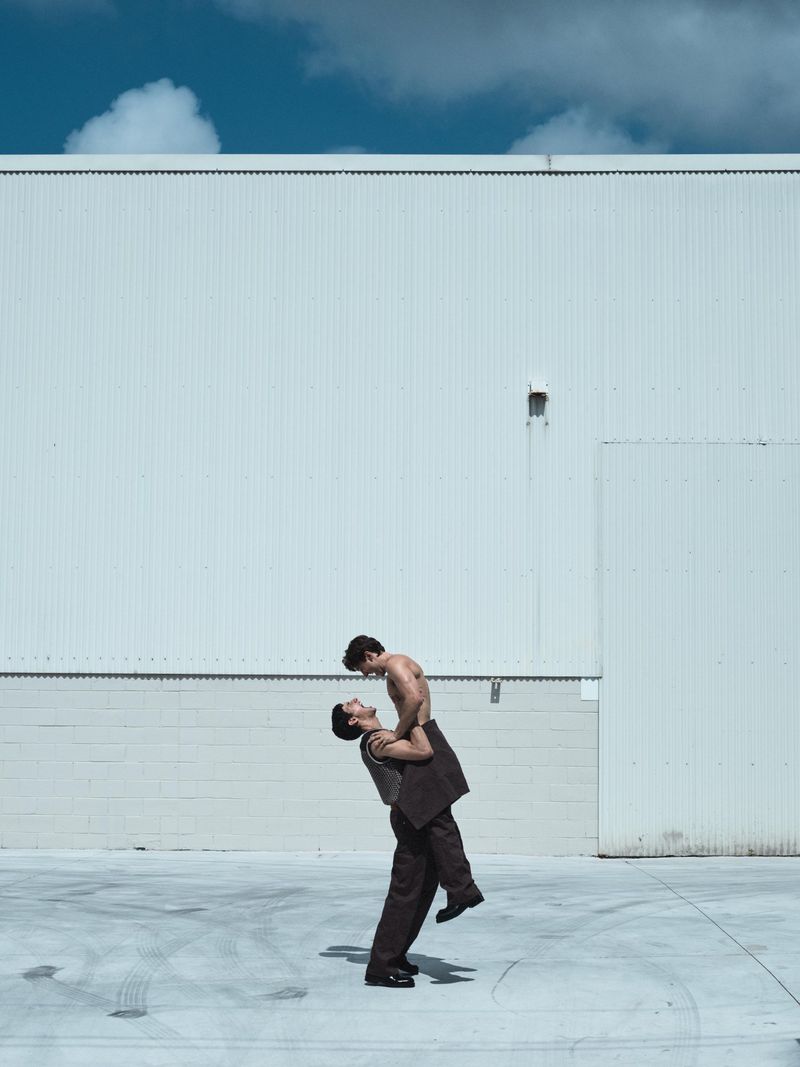 Two men in an outdoor shot: One man in a partial suit holding up another shirtless man against a white building and blue sky.