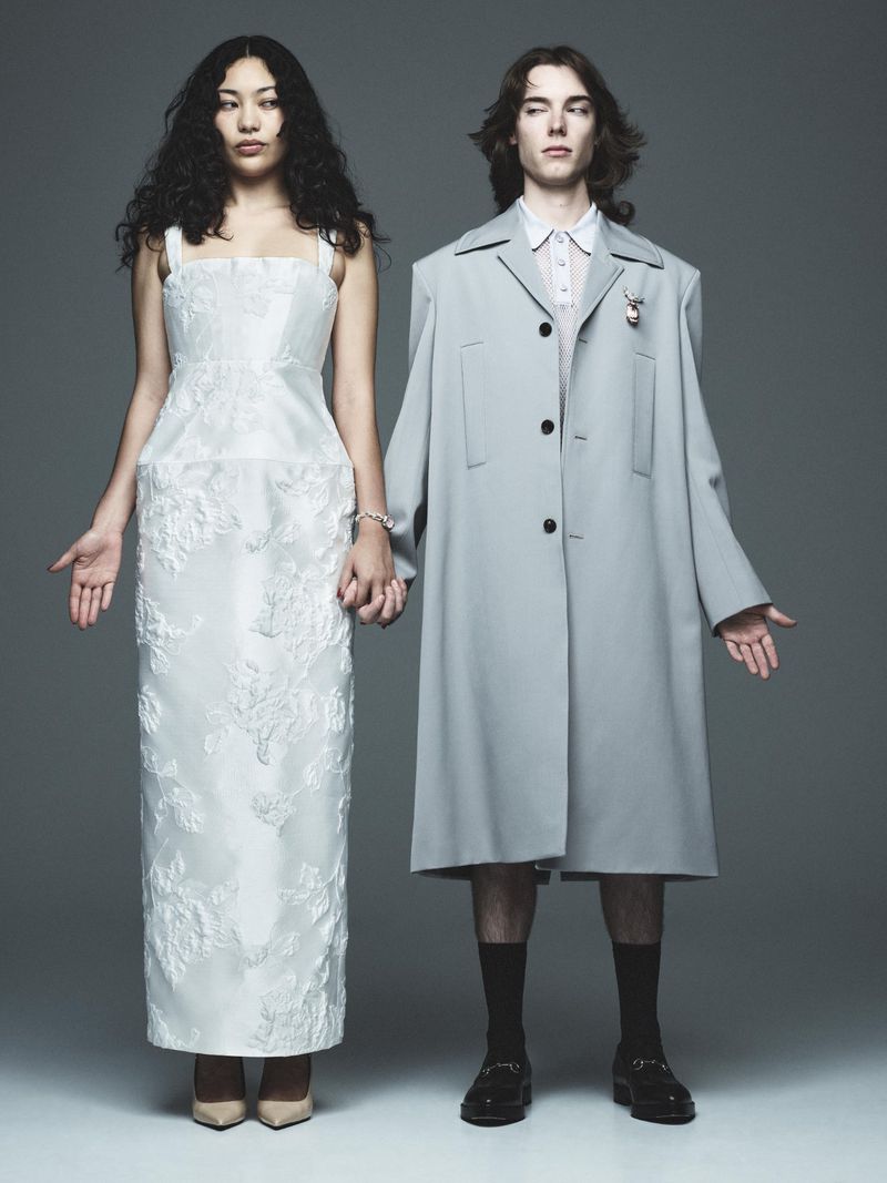 Two fashion models stand together in a studio, holding hands. The woman wears a long white floral dress, and the man wears a grey overcoat.