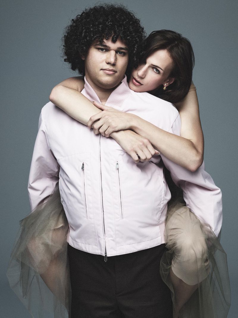 Woman giving a piggyback ride to a man in a photo studio, both wearing light pink jackets and dark pants against a gray backdrop.