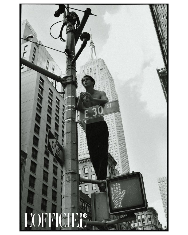 Black and white shot of a shirtless man standing on a traffic light pole with the Empire State Building in the background.