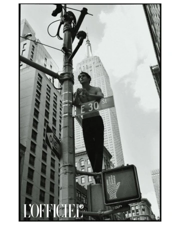 Black and white shot of a shirtless man standing on a traffic light pole with the Empire State Building in the background.