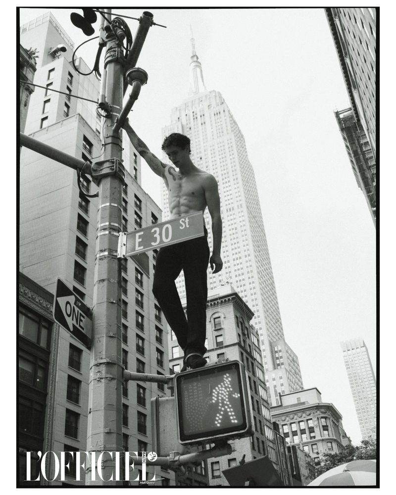 Black and white portrait of a shirtless man standing on a traffic light with the Empire State Building in the background in New York City.